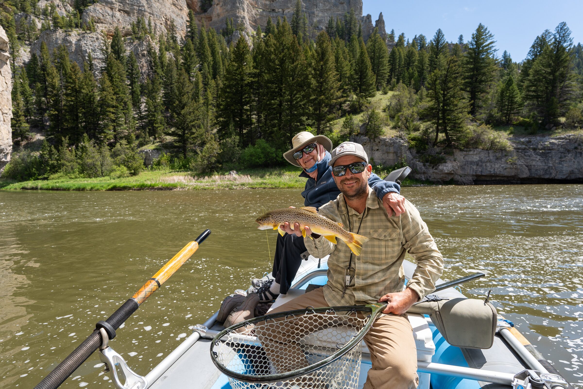 A guide and guest with a brown trout in the raft, the canyon behind them